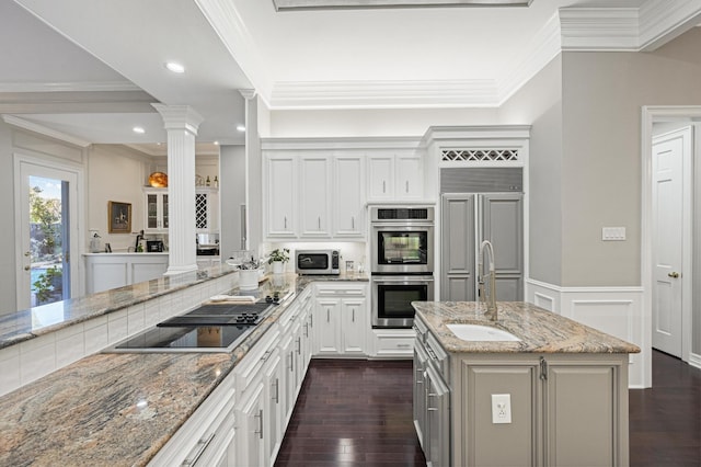 kitchen featuring stainless steel appliances, an island with sink, decorative columns, and light stone counters