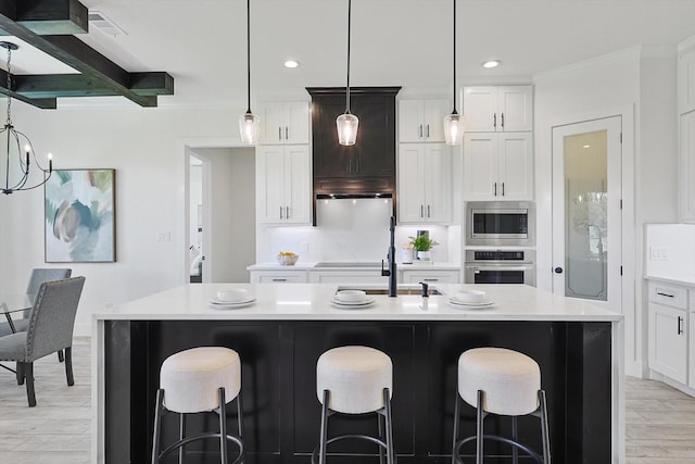 kitchen featuring appliances with stainless steel finishes, decorative light fixtures, beamed ceiling, white cabinets, and a kitchen island with sink