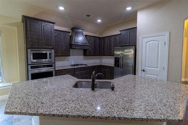 kitchen featuring premium range hood, sink, light stone counters, appliances with stainless steel finishes, and a kitchen island with sink