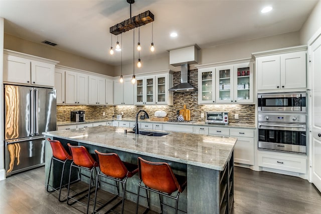 kitchen with wall chimney exhaust hood, stainless steel appliances, sink, and a center island with sink