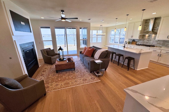 living room featuring light wood finished floors, visible vents, a glass covered fireplace, ceiling fan with notable chandelier, and recessed lighting