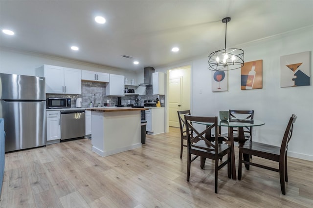 kitchen with appliances with stainless steel finishes, decorative light fixtures, white cabinetry, wall chimney range hood, and light hardwood / wood-style flooring