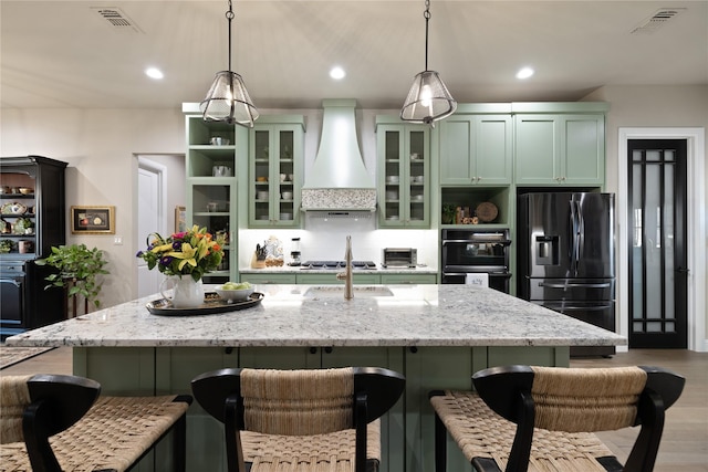 kitchen featuring green cabinets, premium range hood, appliances with stainless steel finishes, light stone countertops, and decorative light fixtures