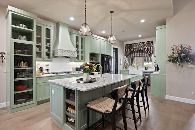 kitchen featuring appliances with stainless steel finishes, custom exhaust hood, a kitchen island with sink, green cabinets, and light stone counters