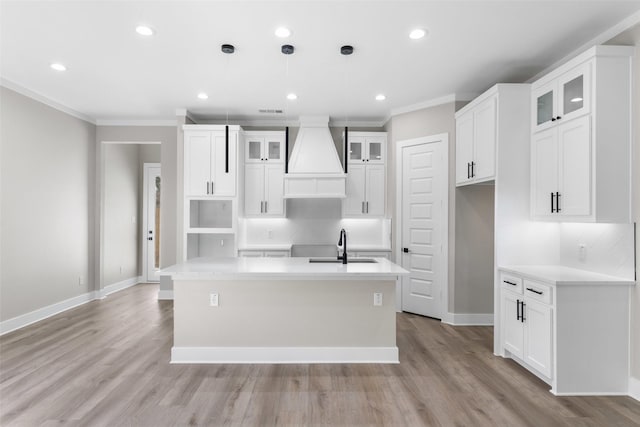 kitchen featuring crown molding, a center island with sink, custom range hood, light wood-style floors, and a sink