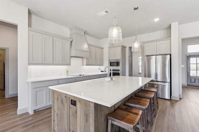 kitchen featuring sink, custom exhaust hood, appliances with stainless steel finishes, an island with sink, and pendant lighting