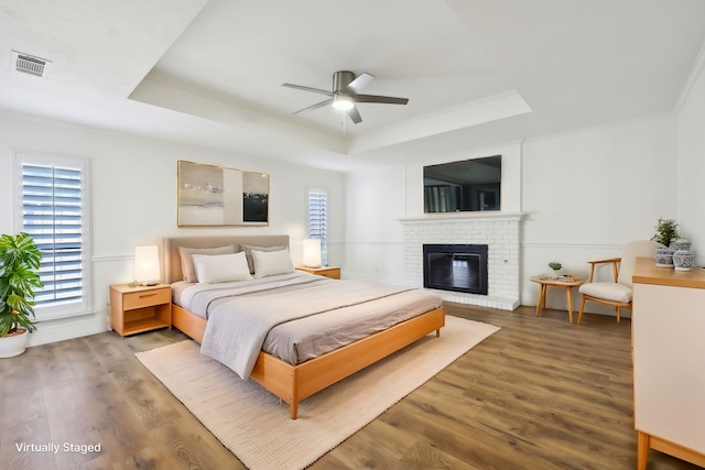 bedroom with crown molding, ceiling fan, a tray ceiling, wood-type flooring, and a brick fireplace