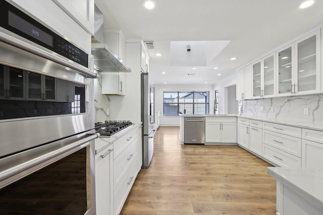 kitchen featuring wall chimney range hood, light hardwood / wood-style flooring, appliances with stainless steel finishes, light stone countertops, and white cabinets