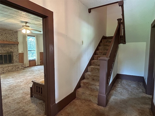 stairway with a fireplace, ceiling fan, and carpet