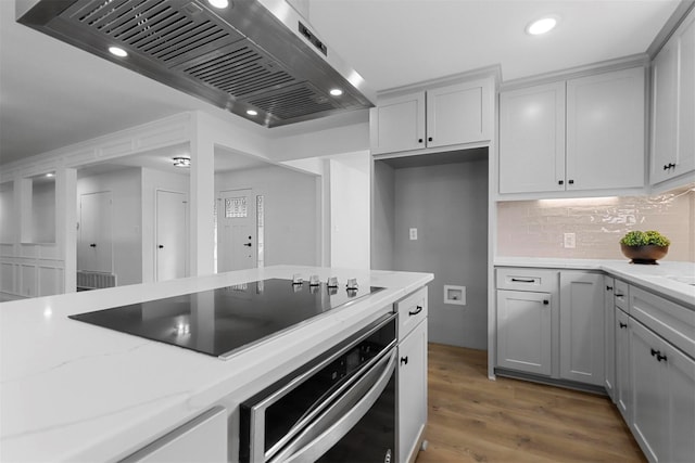 kitchen featuring black electric cooktop, stainless steel oven, ventilation hood, light wood-type flooring, and light stone countertops