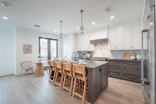 kitchen with white cabinetry, dark brown cabinets, an island with sink, custom range hood, and pendant lighting