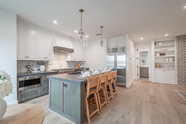 kitchen featuring appliances with stainless steel finishes, white cabinetry, a kitchen island with sink, custom range hood, and decorative light fixtures