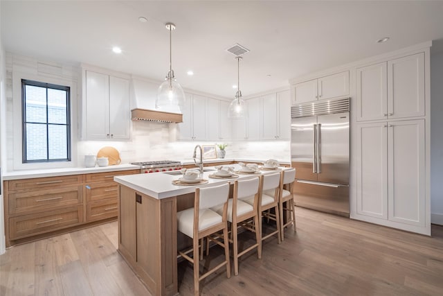 kitchen with appliances with stainless steel finishes, white cabinetry, light hardwood / wood-style floors, an island with sink, and decorative light fixtures