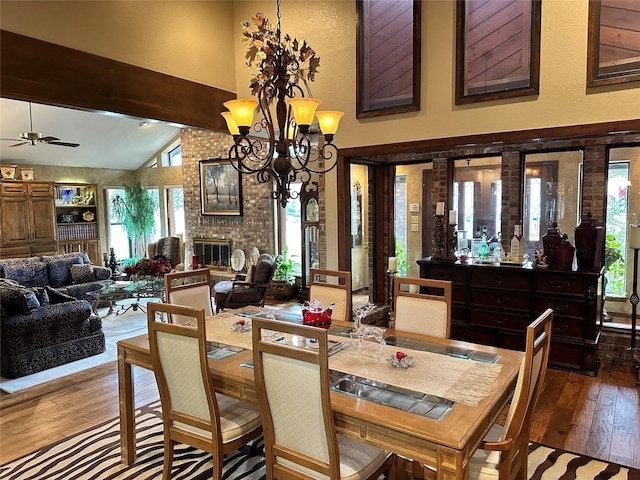 dining space with ceiling fan with notable chandelier, high vaulted ceiling, hardwood / wood-style flooring, a brick fireplace, and beam ceiling