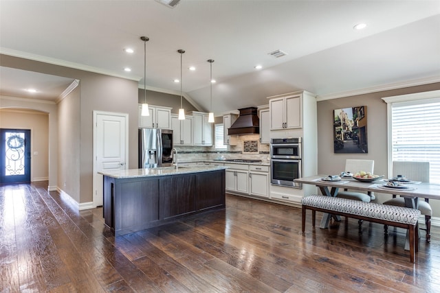 kitchen featuring lofted ceiling, backsplash, a kitchen island with sink, stainless steel appliances, and custom range hood