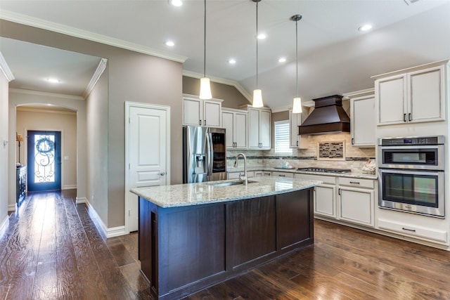 kitchen featuring premium range hood, sink, stainless steel appliances, light stone countertops, and a kitchen island with sink