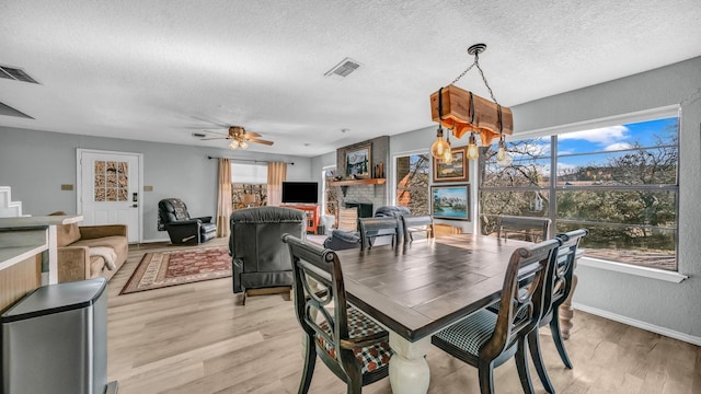 dining room featuring a stone fireplace, ceiling fan with notable chandelier, a textured ceiling, and light wood-type flooring