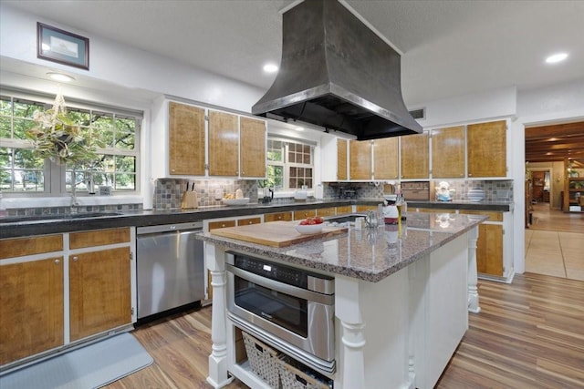 kitchen with tasteful backsplash, appliances with stainless steel finishes, hardwood / wood-style flooring, island exhaust hood, and a kitchen island with sink