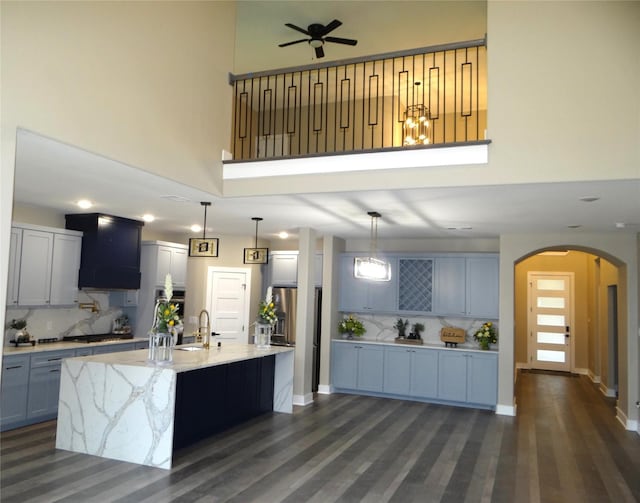 kitchen featuring backsplash, hanging light fixtures, a towering ceiling, light stone countertops, and an island with sink