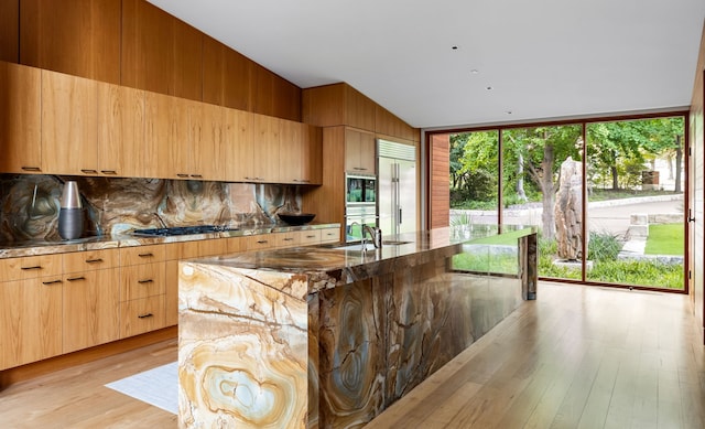 kitchen with sink, built in appliances, a center island with sink, stone counters, and light hardwood / wood-style floors