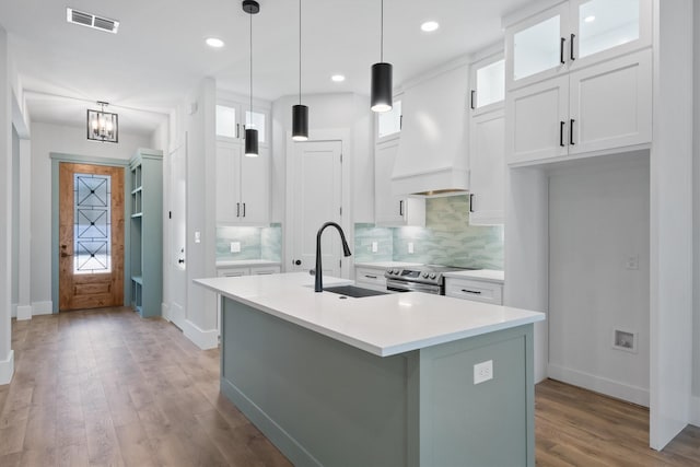 kitchen with stainless steel electric stove, premium range hood, sink, white cabinets, and a center island with sink