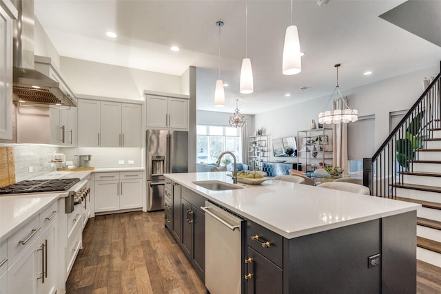 kitchen with wall chimney range hood, sink, a kitchen island with sink, hanging light fixtures, and stainless steel appliances