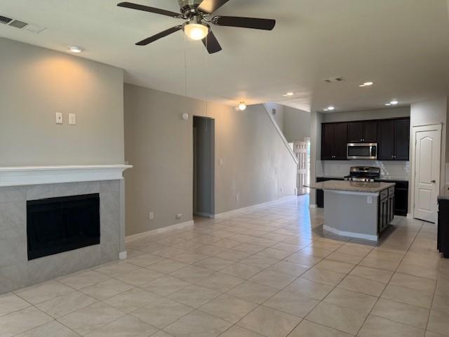 kitchen featuring light tile patterned flooring, appliances with stainless steel finishes, an island with sink, ceiling fan, and a tiled fireplace