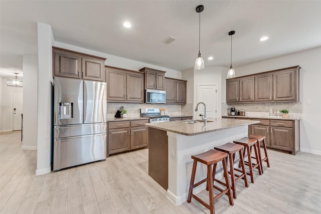 kitchen with an island with sink, sink, light stone counters, stainless steel appliances, and light wood-type flooring