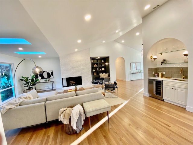 living room featuring high vaulted ceiling, wine cooler, light hardwood / wood-style floors, a brick fireplace, and wet bar