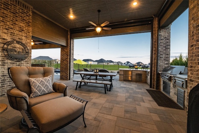 patio terrace at dusk featuring ceiling fan, an outdoor kitchen, and a grill