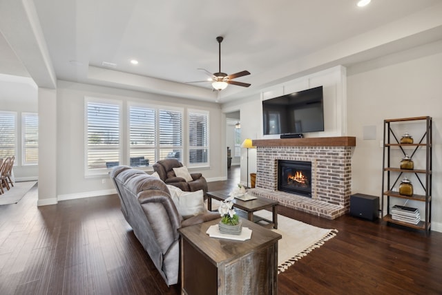 living room with dark wood-type flooring, ceiling fan, a raised ceiling, and a brick fireplace