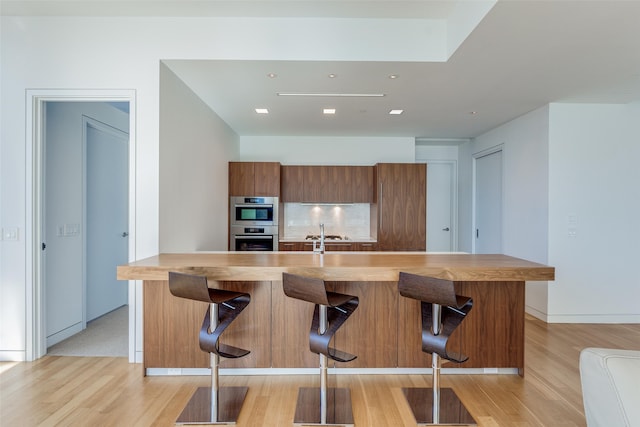 kitchen with double oven, a breakfast bar, a kitchen island with sink, and light wood-type flooring