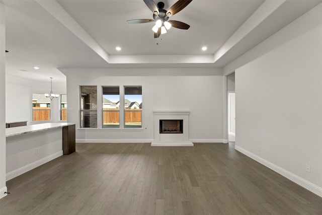 unfurnished living room with ceiling fan with notable chandelier, dark wood-type flooring, and a raised ceiling