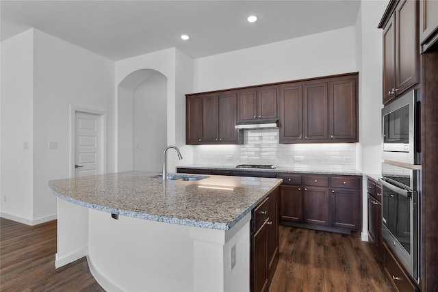 kitchen with sink, dark wood-type flooring, appliances with stainless steel finishes, light stone countertops, and a center island with sink