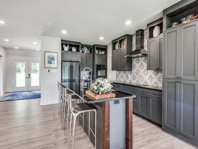 kitchen with french doors, a kitchen breakfast bar, wall chimney range hood, a kitchen island with sink, and black appliances