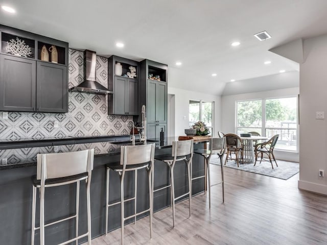 kitchen with lofted ceiling, a breakfast bar area, light hardwood / wood-style flooring, decorative backsplash, and wall chimney range hood