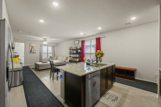 kitchen featuring a center island with sink, stainless steel appliances, light stone counters, dark brown cabinetry, and a kitchen bar