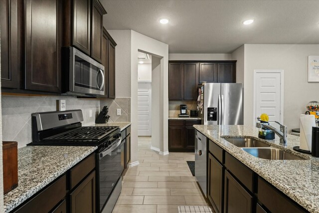 kitchen featuring dark brown cabinetry, sink, light stone counters, appliances with stainless steel finishes, and a kitchen island with sink