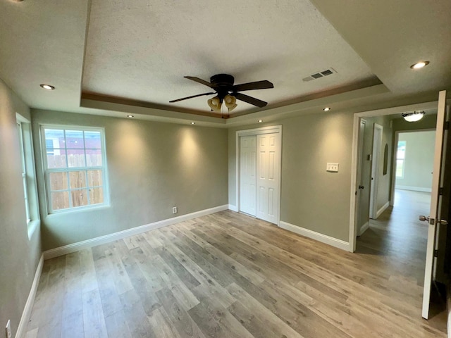 unfurnished bedroom featuring ceiling fan, a tray ceiling, a textured ceiling, a closet, and light wood-type flooring