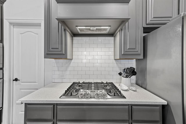 kitchen featuring gray cabinets, backsplash, light stone counters, stainless steel appliances, and wall chimney exhaust hood