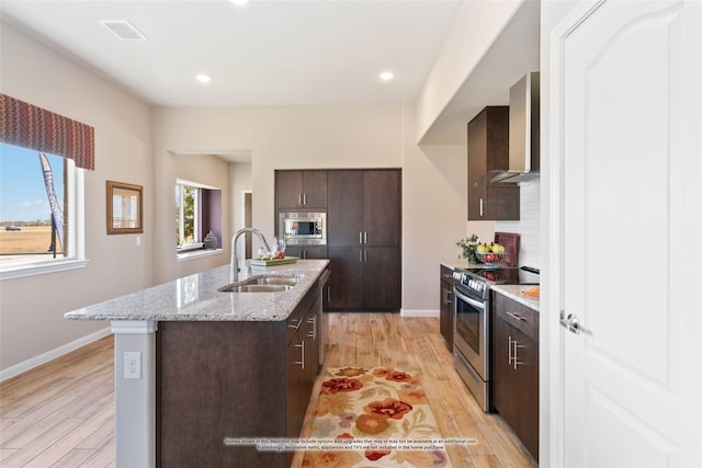 kitchen with sink, appliances with stainless steel finishes, a kitchen island with sink, light hardwood / wood-style floors, and wall chimney exhaust hood