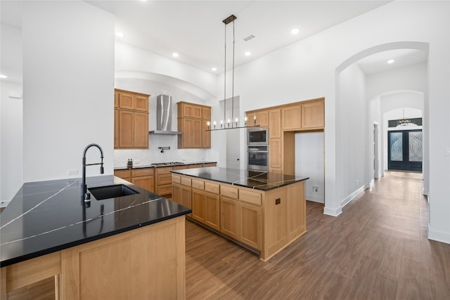 kitchen with appliances with stainless steel finishes, a towering ceiling, sink, a kitchen island with sink, and wall chimney range hood