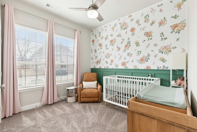 carpeted bedroom featuring a crib, wallpapered walls, multiple windows, and a wainscoted wall