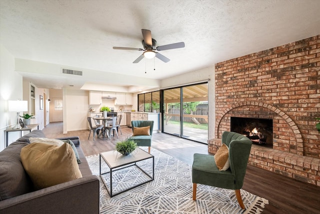 living room with ceiling fan, a brick fireplace, a textured ceiling, and light hardwood / wood-style flooring