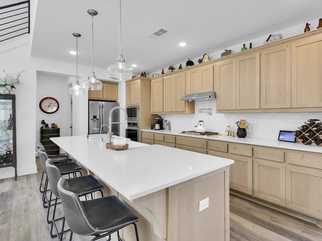 kitchen featuring visible vents, light brown cabinetry, under cabinet range hood, appliances with stainless steel finishes, and a kitchen island with sink