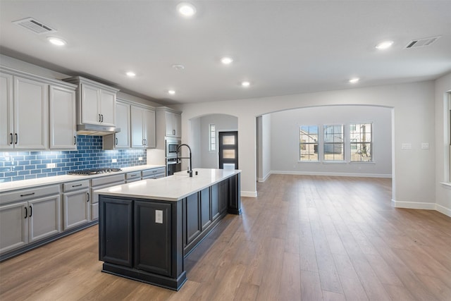 kitchen with gray cabinetry, backsplash, light hardwood / wood-style flooring, and a center island with sink