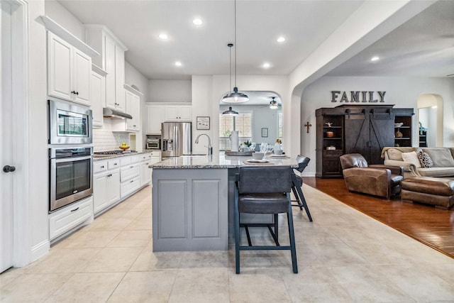 kitchen with appliances with stainless steel finishes, hanging light fixtures, an island with sink, light stone counters, and white cabinets