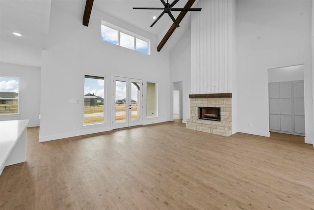 unfurnished living room with light hardwood / wood-style flooring, ceiling fan, a stone fireplace, french doors, and beamed ceiling