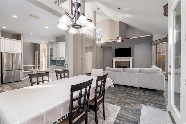 dining area featuring dark wood finished floors, a fireplace, recessed lighting, high vaulted ceiling, and ceiling fan with notable chandelier