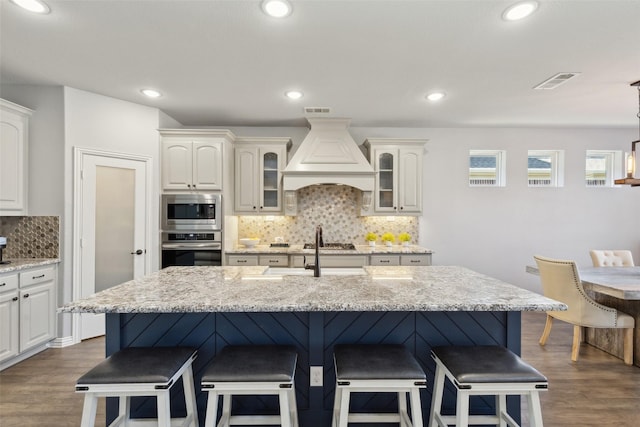 kitchen featuring premium range hood, backsplash, a kitchen island with sink, light stone counters, and stainless steel appliances